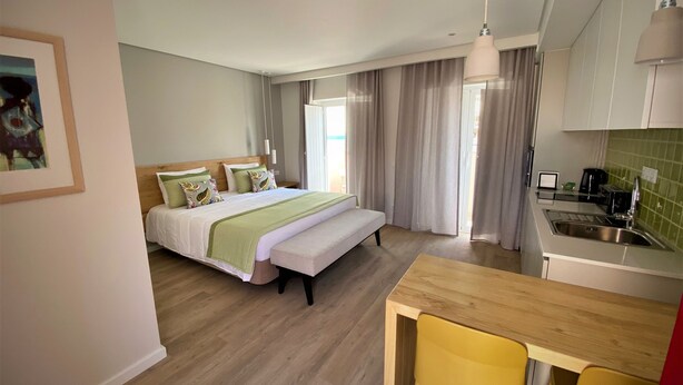 A modern hotel room with a bed, a wooden desk, a yellow chair, and a kitchenette. - Cardeal Suites & Apartments (Faro)