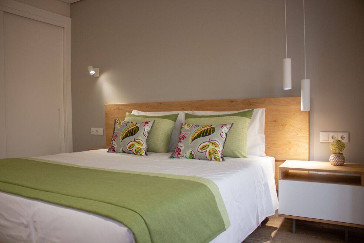 A neatly made bed with a green bedspread and floral pillows, a wooden headboard, a bedside table with a vase, and a wall-mounted light fixture.