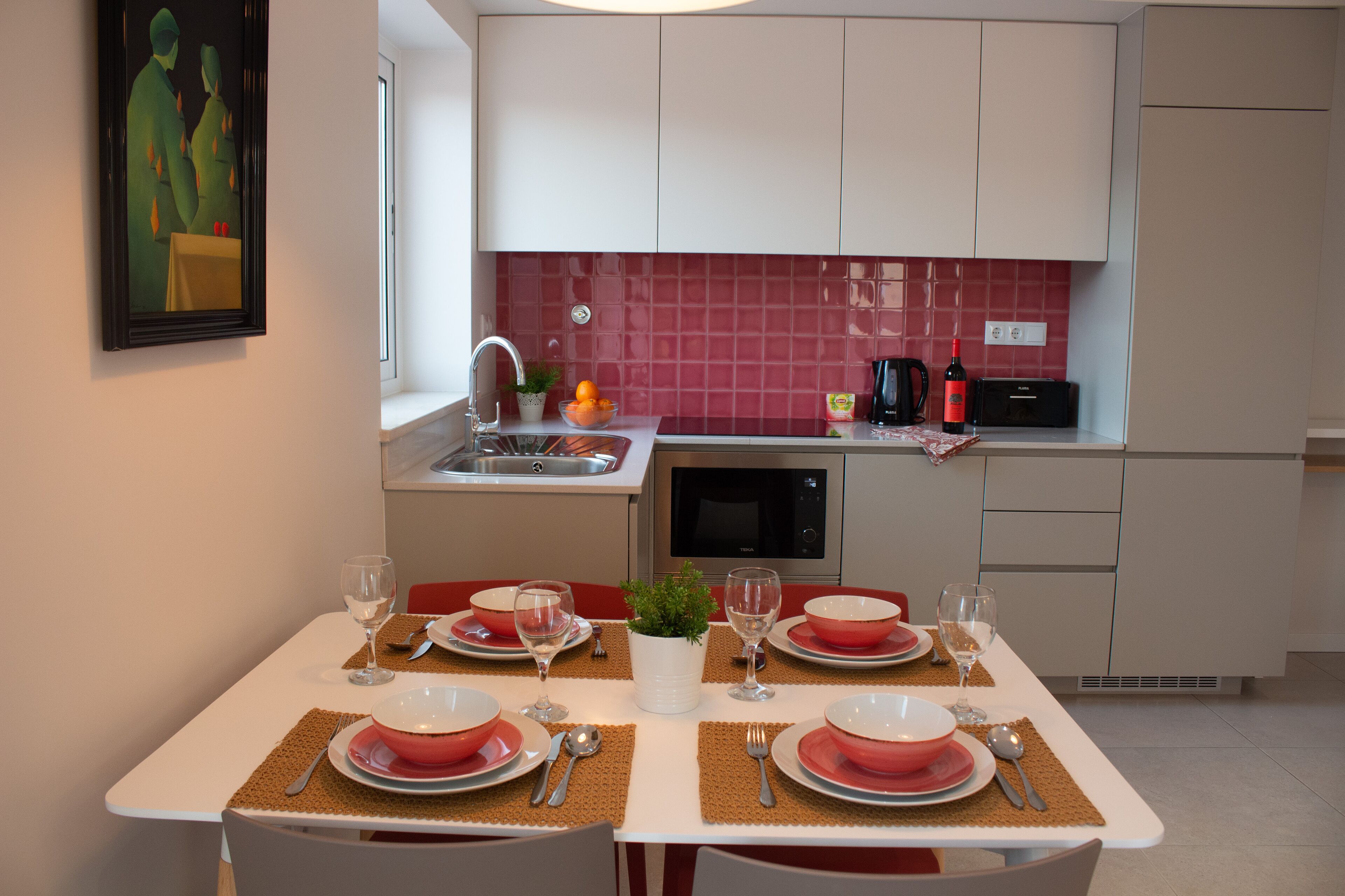 A modern kitchen with white cabinets, a red backsplash, and a dining area set for four with red plates and silverware.