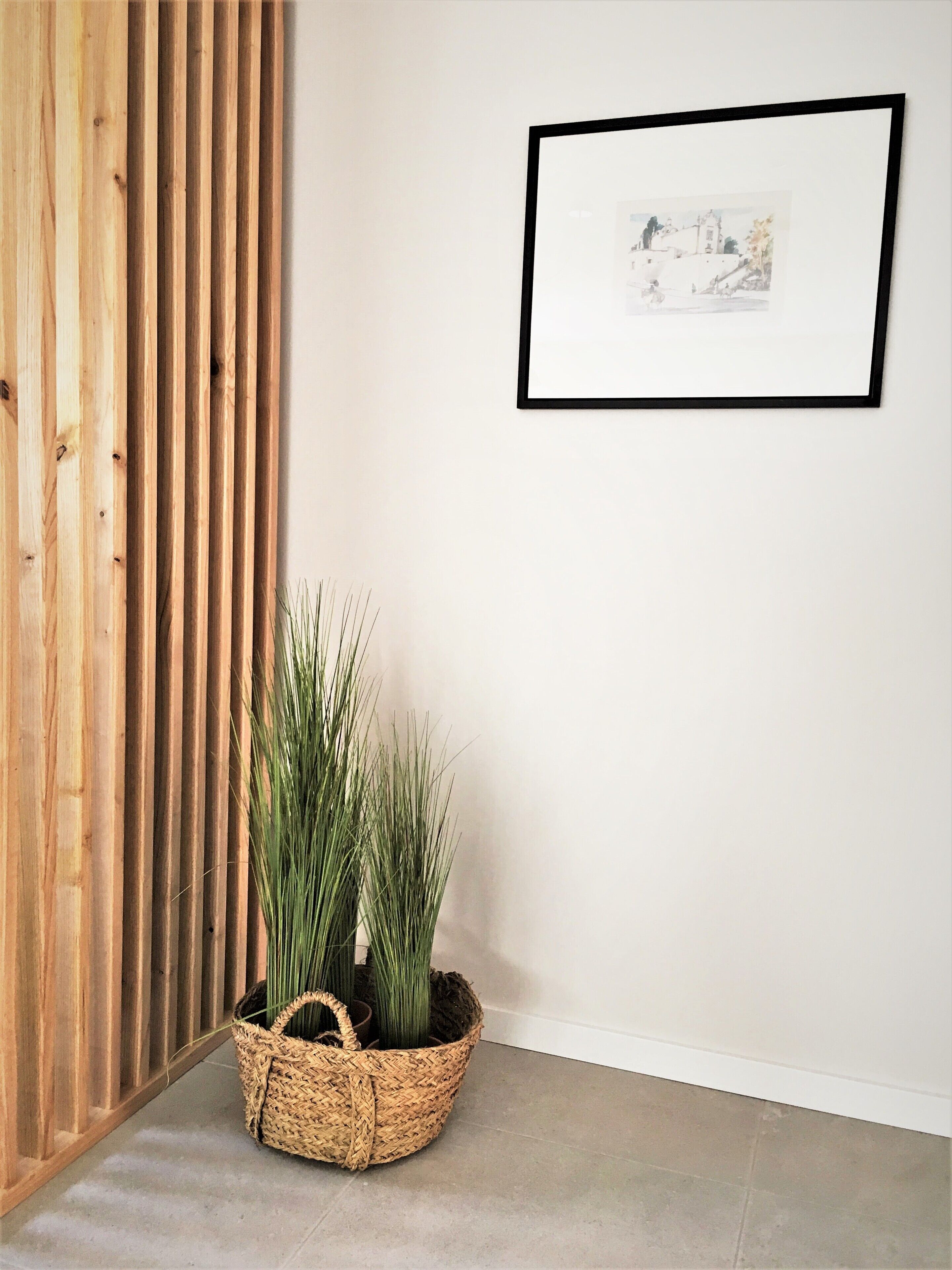 A corner of a room with a wooden slat wall, a framed picture on the wall, and a woven basket with plants on the floor.
