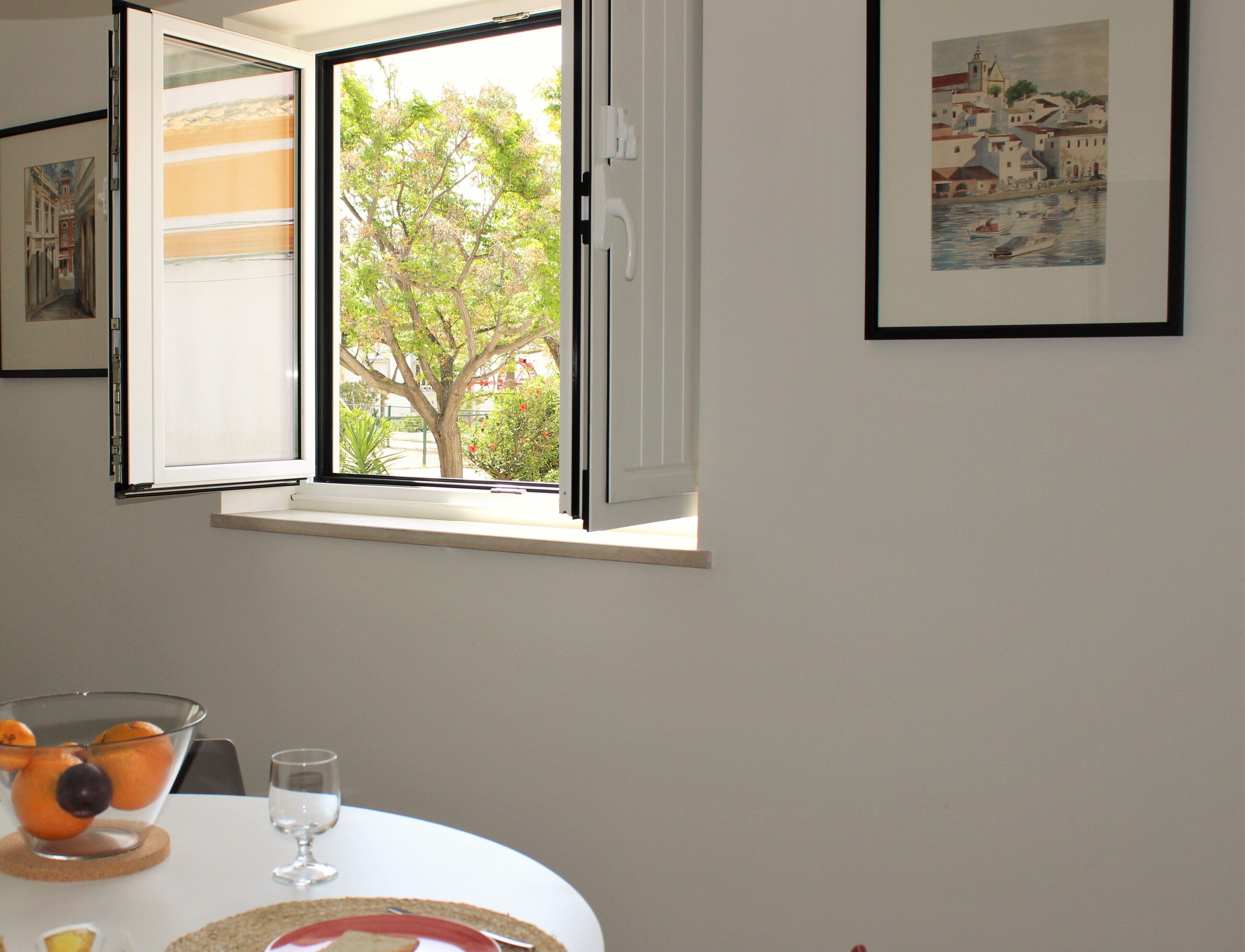 Un coin repas avec une table dressée pour deux personnes, une fenêtre dont le volet est entrouvert et des œuvres d’art encadrées accrochées au mur.