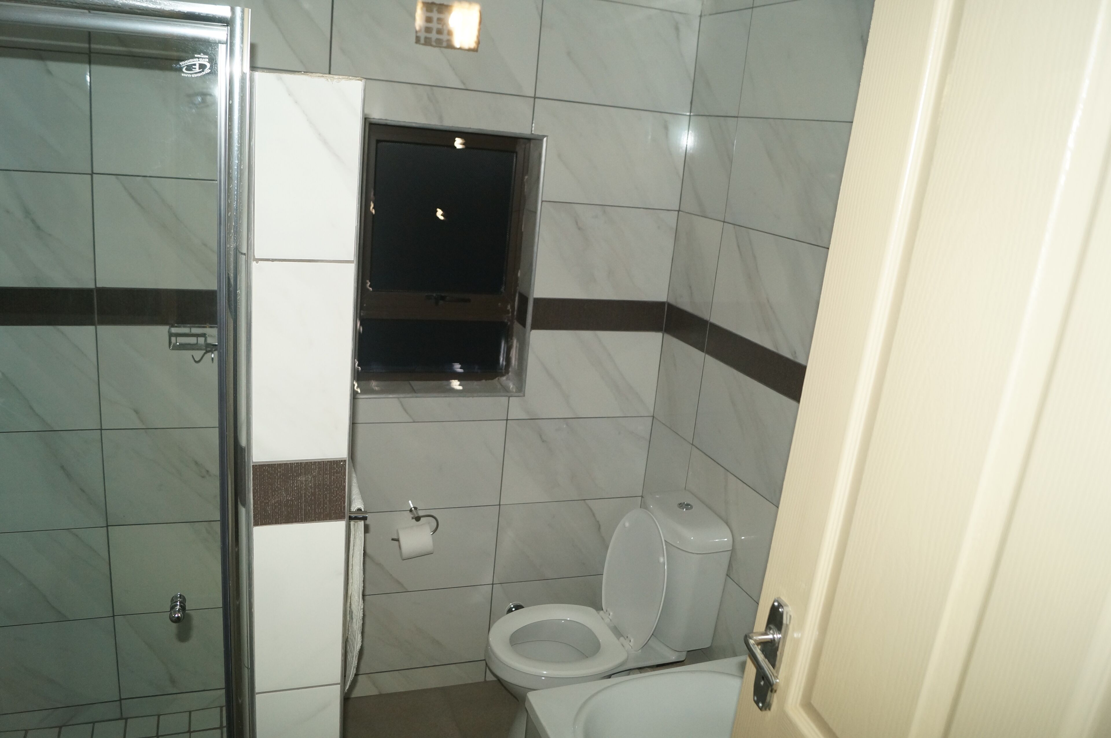 Een badkamer met een douchecabine van glas, een toilet en een klein raam boven het toilet.