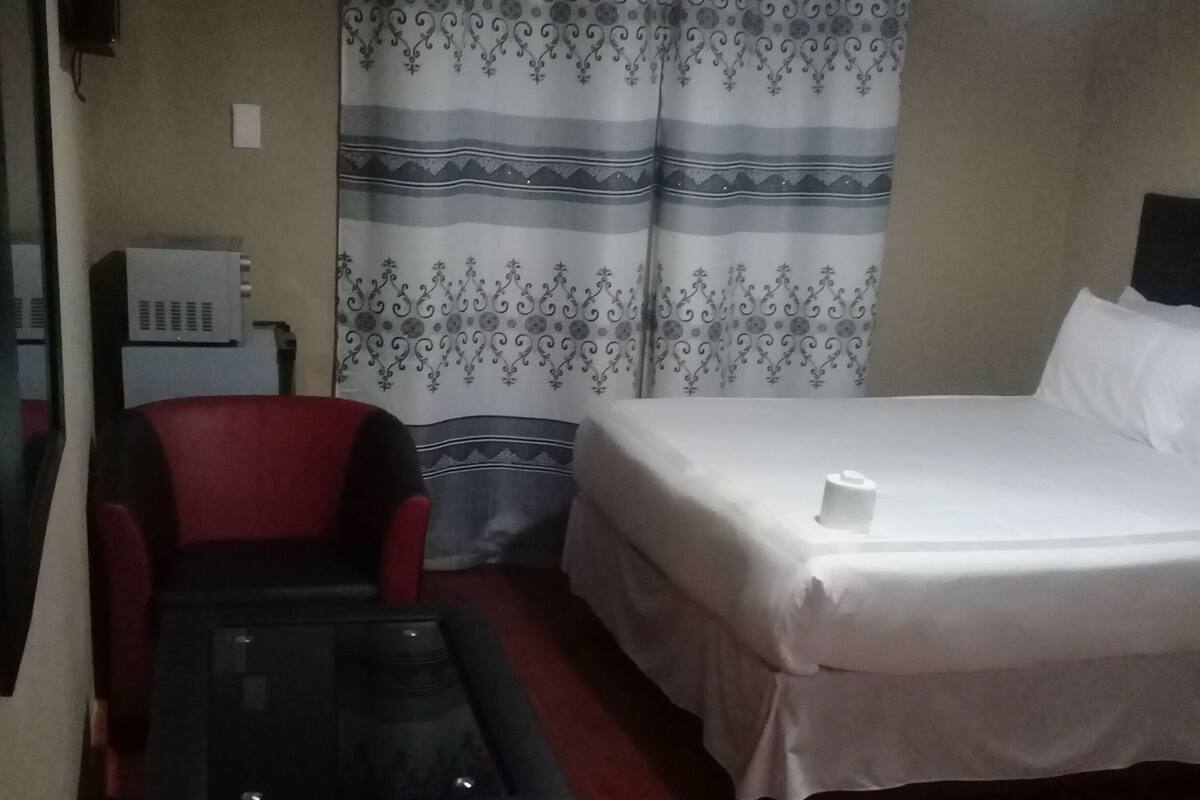 Une pièce comprenant un lit, une petite table et une chaise. On y trouve un rideau à motifs, un radiateur et un téléviseur fixé au mur.