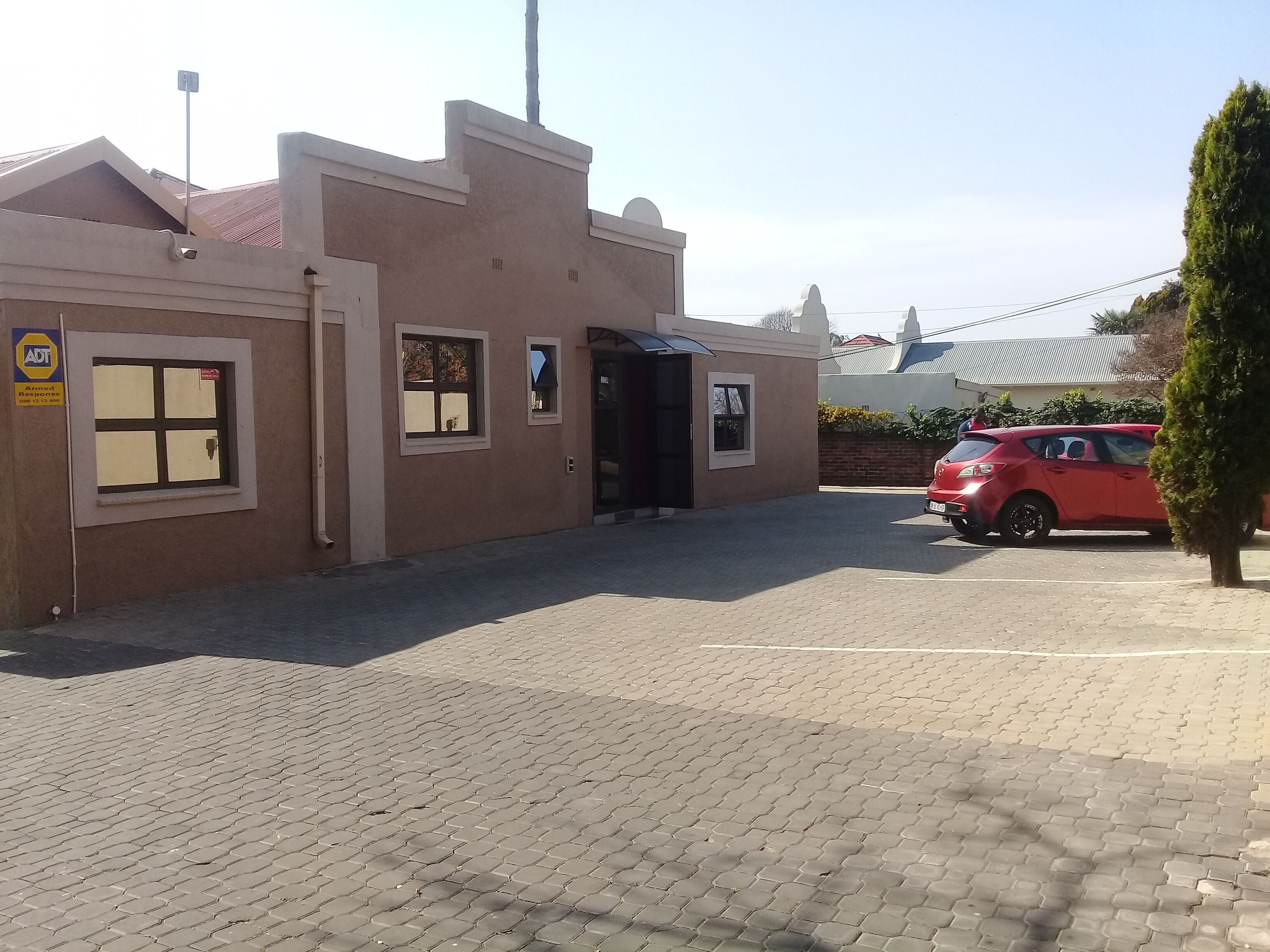 A building with a paved entrance, a red car parked in front, and a small tree to the right.
