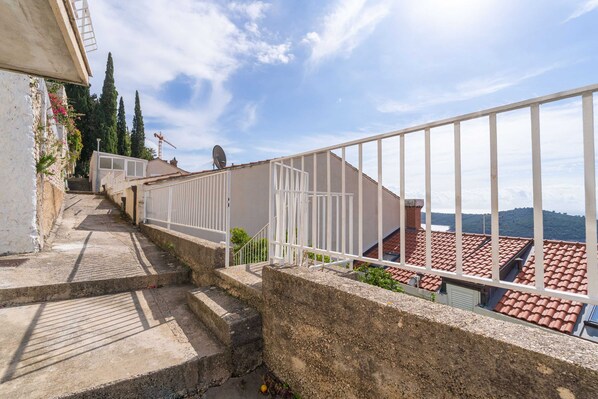 A concrete staircase leading to a terrace with white railings, a satellite dish, and a view of red-tiled roofs and distant hills. - Apartments Cicko (Dubrovnik)