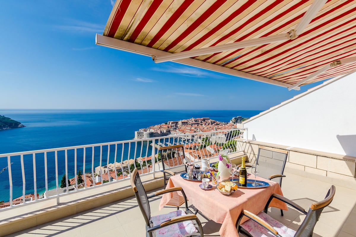 A balcony with a table set for dining, overlooking a coastal town and the sea.