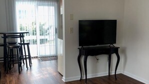 A living room with a TV on a stand, wooden flooring, and a dining area with chairs.