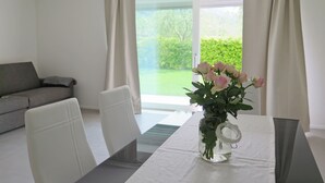 A modern living room with a sofa, dining table, and chairs. A vase with flowers is on the table. - 32 GARDEN HOUSE (Nago-Torbole)