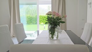 A dining table with a vase of flowers, white chairs, and a view of a garden through a glass door. - 32 GARDEN HOUSE (Nago-Torbole)