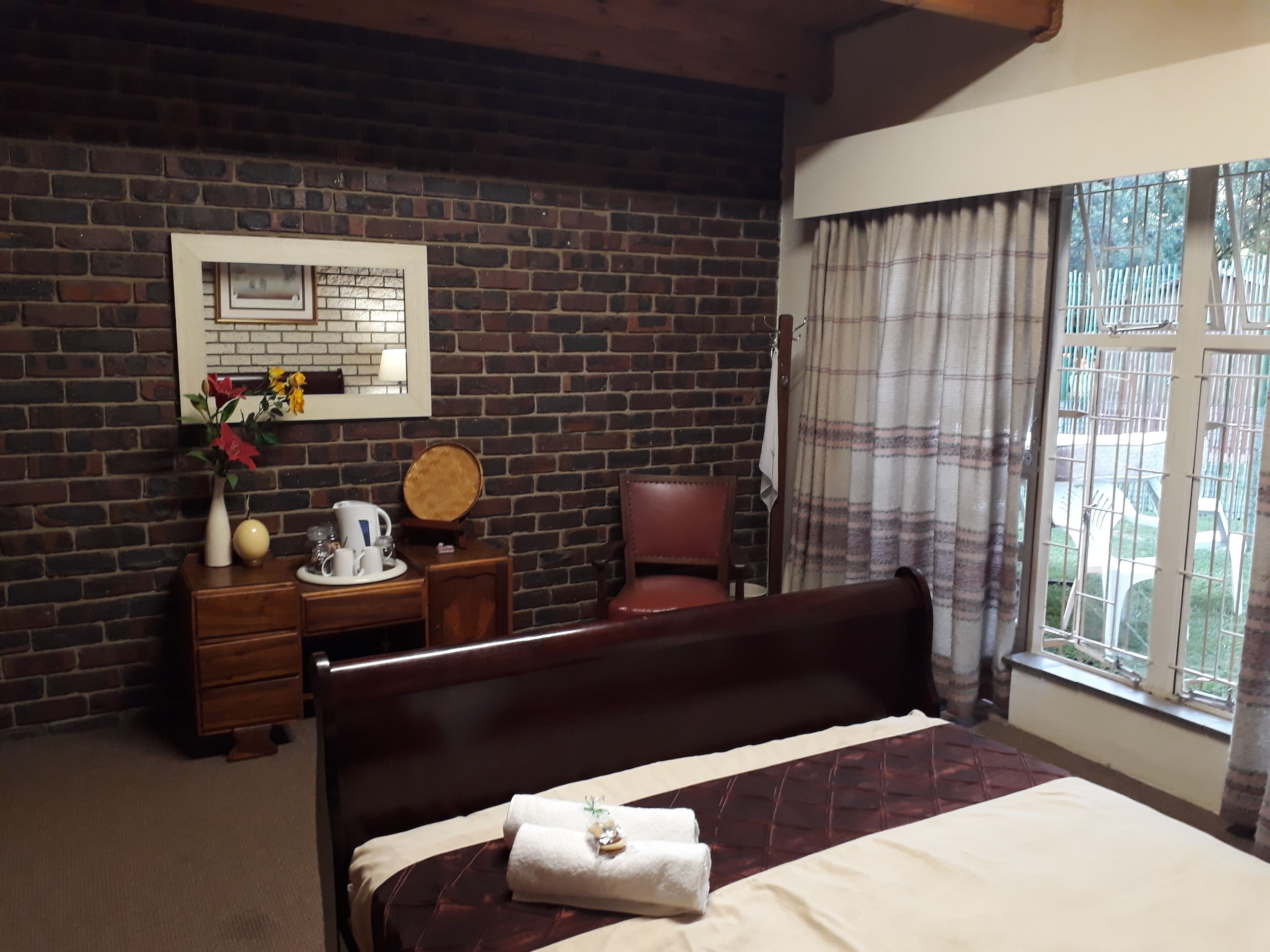 Habitación de hotel con pared de ladrillo, cama de madera, escritorio con silla, ventana con cortinas y espejo en la pared.