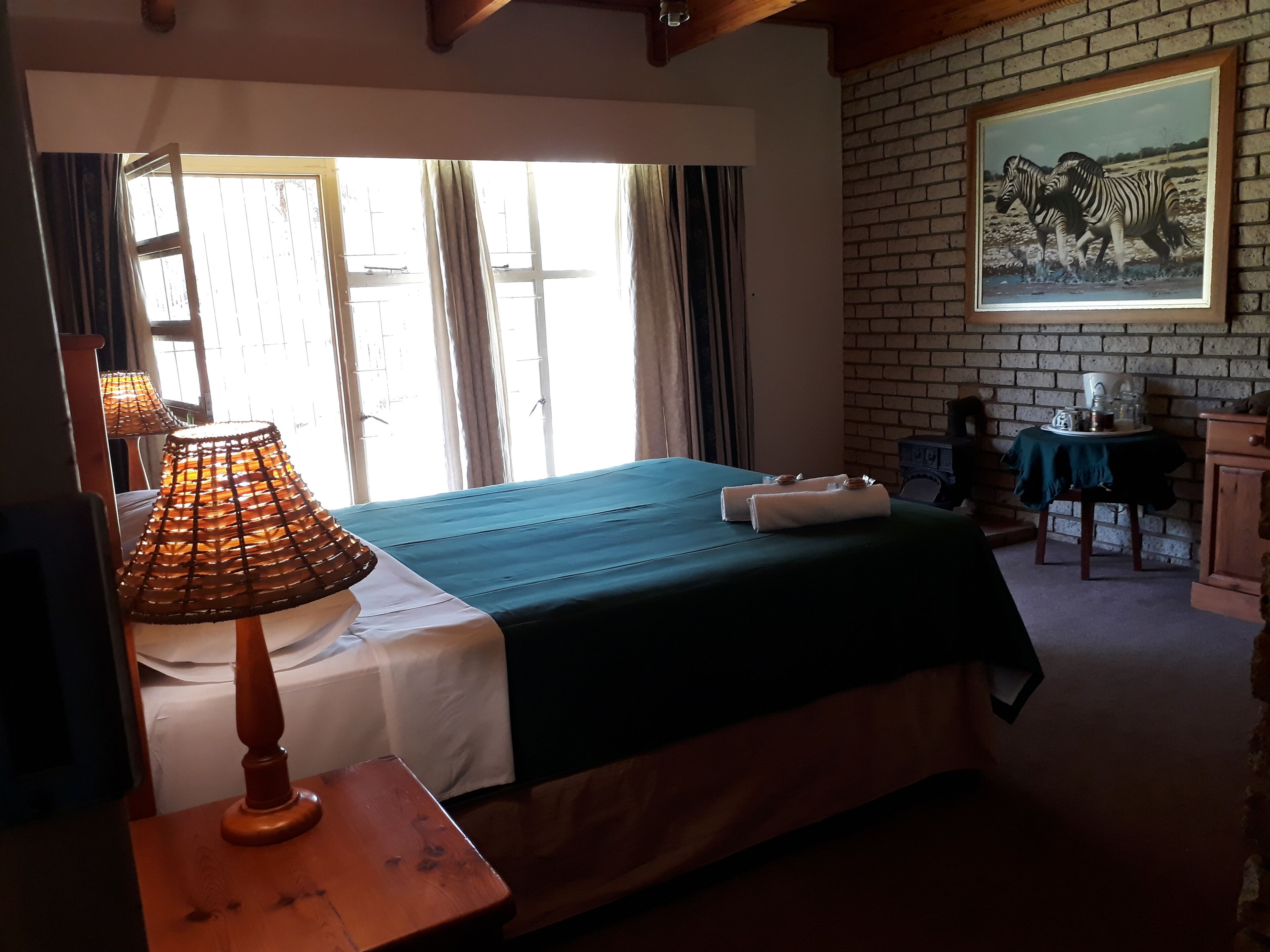 A room with a bed, a table with a lamp, a painting on the wall, and a window with curtains.