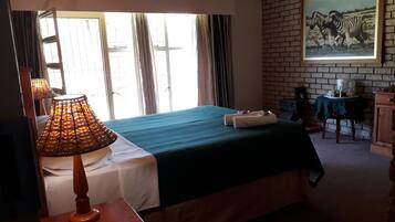 A room with a bed, a table with a lamp, a painting on the wall, and a window with curtains.