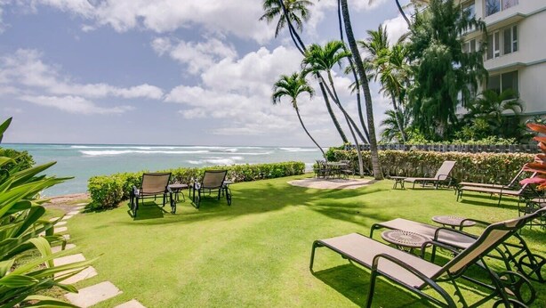 A well-maintained outdoor area with a lawn, lounge chairs, and tables, overlooking a beach with palm trees and a clear sky.