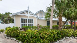 A two-story house with a beige exterior, white trim, and a peaked roof. It is surrounded by a landscaped garden with various shrubs and a palm tree.