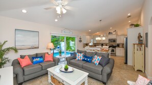 A spacious living room with a grey sofa set, a coffee table, and a ceiling fan. A kitchen is visible in the background with white cabinets and a dining area.