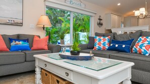 A living room with two grey sofas, colorful pillows, a white coffee table, a lamp, and a window with a view of greenery.