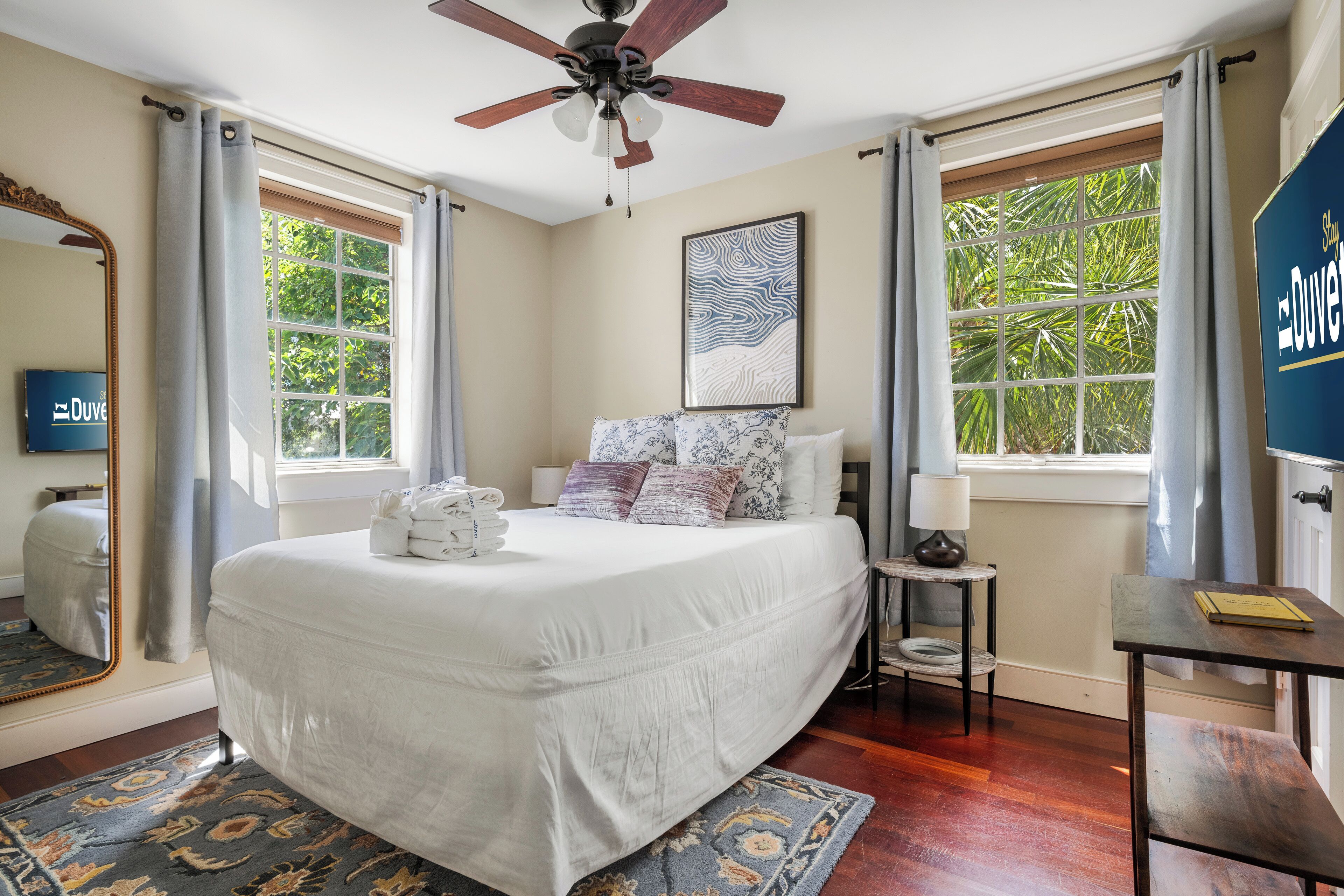 A bedroom with a bed, a ceiling fan, a window with curtains, a bedside table with a lamp, and a framed picture on the wall.