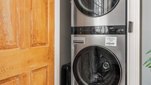 A compact, stainless steel washer and dryer unit with a Duet label, positioned in a hallway with wooden flooring and a white door. - Easy Walk to King Street! | Casa Fuego Suite E (Charleston)