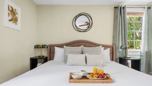 A neatly made bed with a tray of breakfast, a bedside table with a lamp, and a window with curtains. - Easy Walk to King Street! | Casa Fuego Suite E (Charleston)