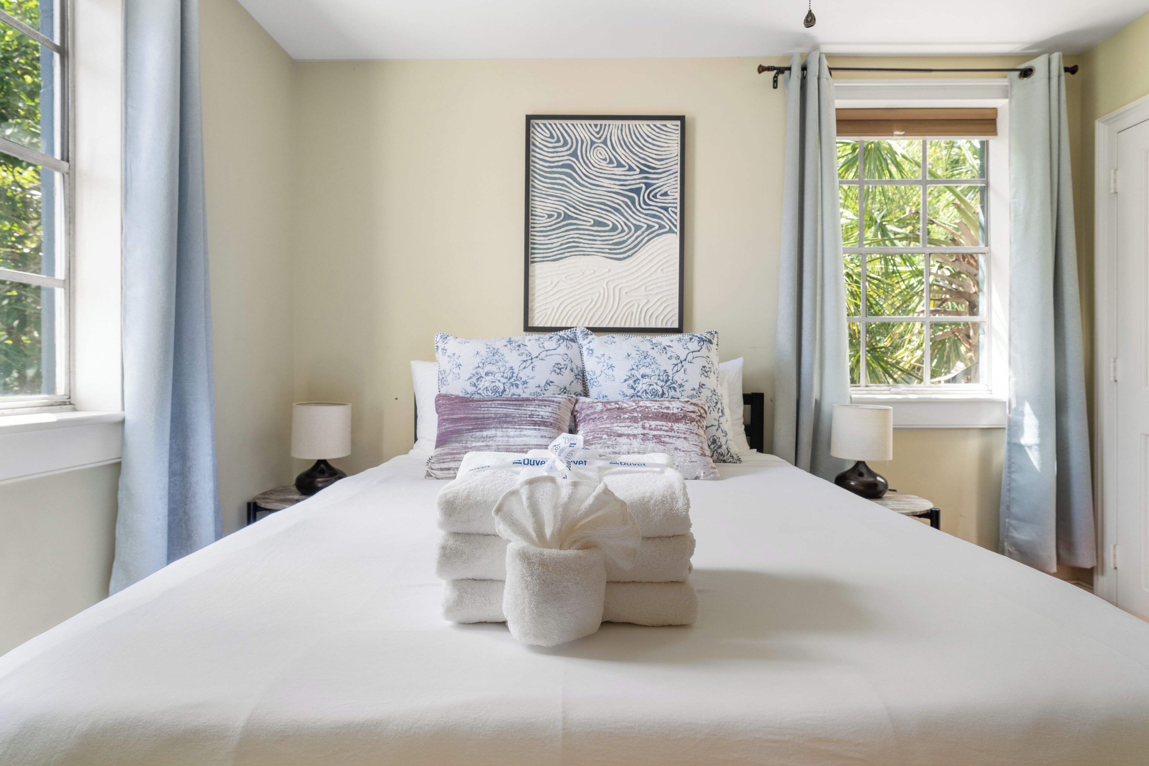 A neatly made bed with towels, a framed artwork on the wall, and a window with curtains.