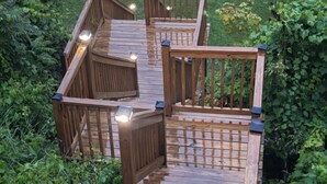 A wooden deck with railings and lights, surrounded by greenery.