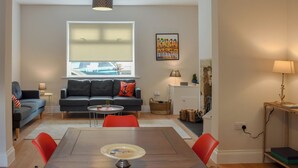 A living room with a sofa, chairs, a dining table, a lamp, a window with blinds, a painting, and a small table with a lamp. - 3 bedroom accommodation in Amble, near Morpeth (Amble, near Morpeth)