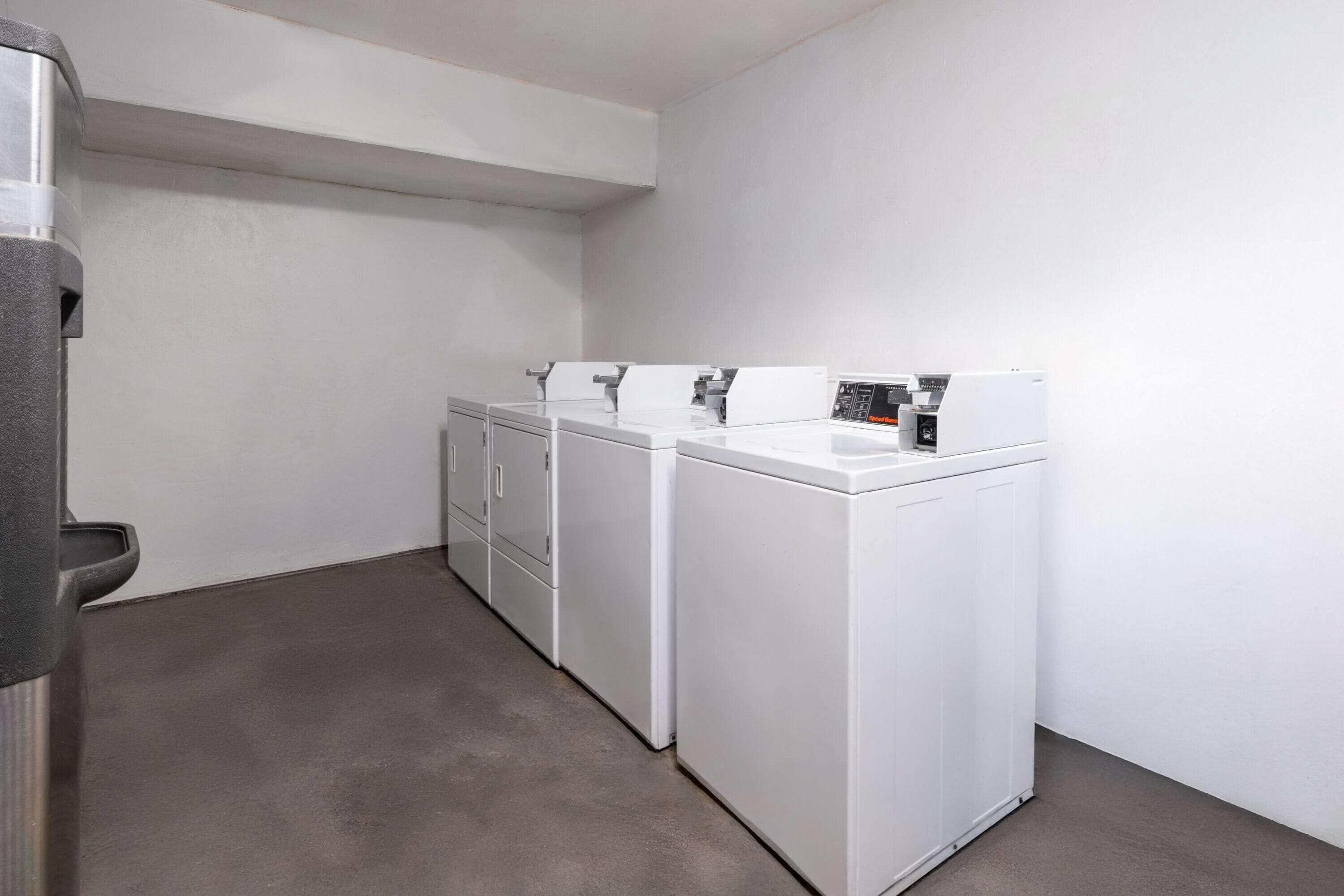 Una sala de lavandería con tres lavadoras y secadoras blancas, una secadora de metal y un piso gris.