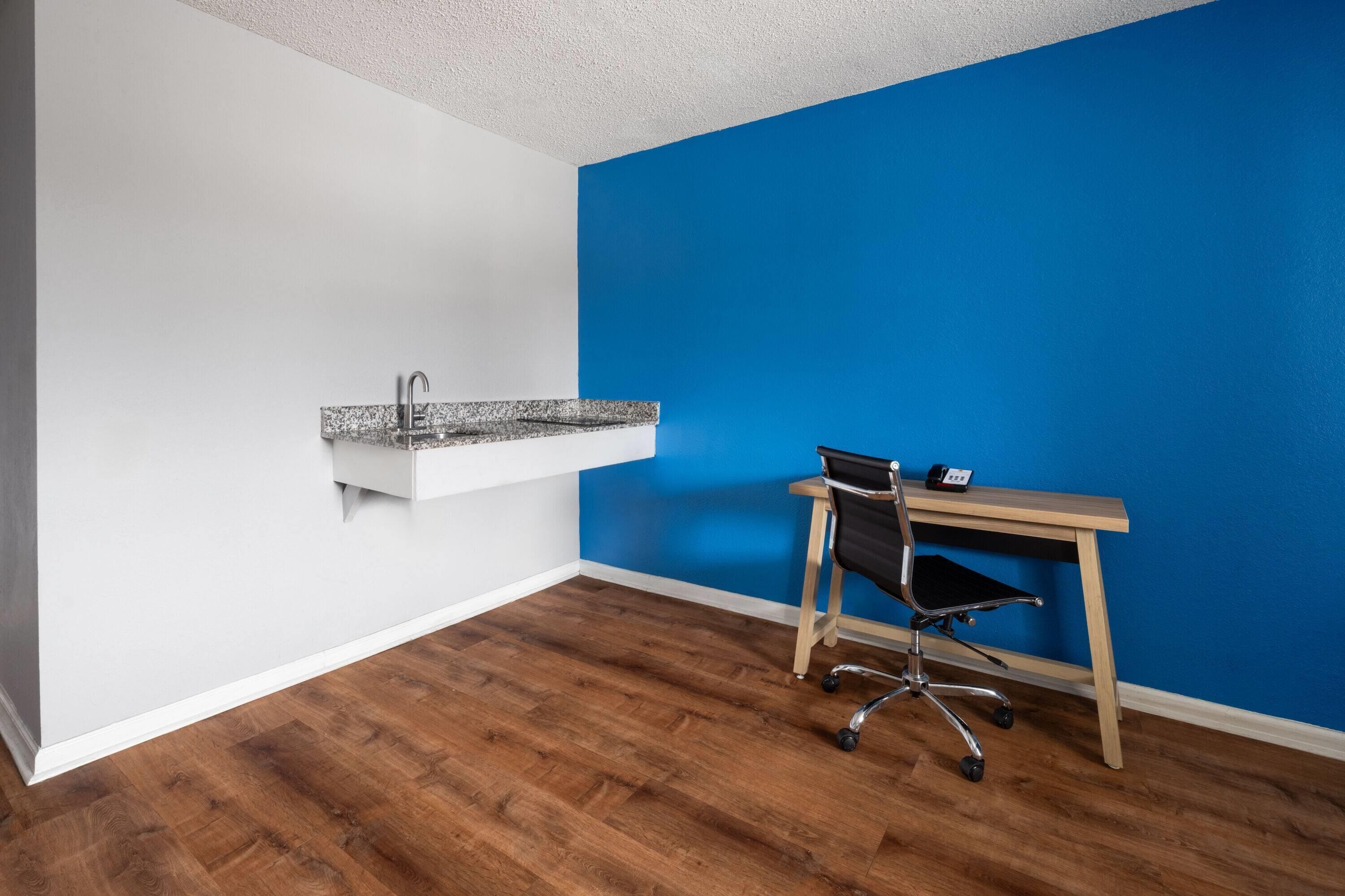 A room with a wooden floor, a white wall, a blue accent wall, a built-in kitchen sink, and a desk with a chair.