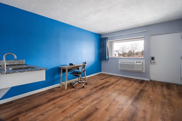 A room with a blue accent wall, wooden flooring, a desk with a chair, a window with a view, and a door. - Super 8 by Wyndham Portsmouth - Norfolk (Portsmouth)