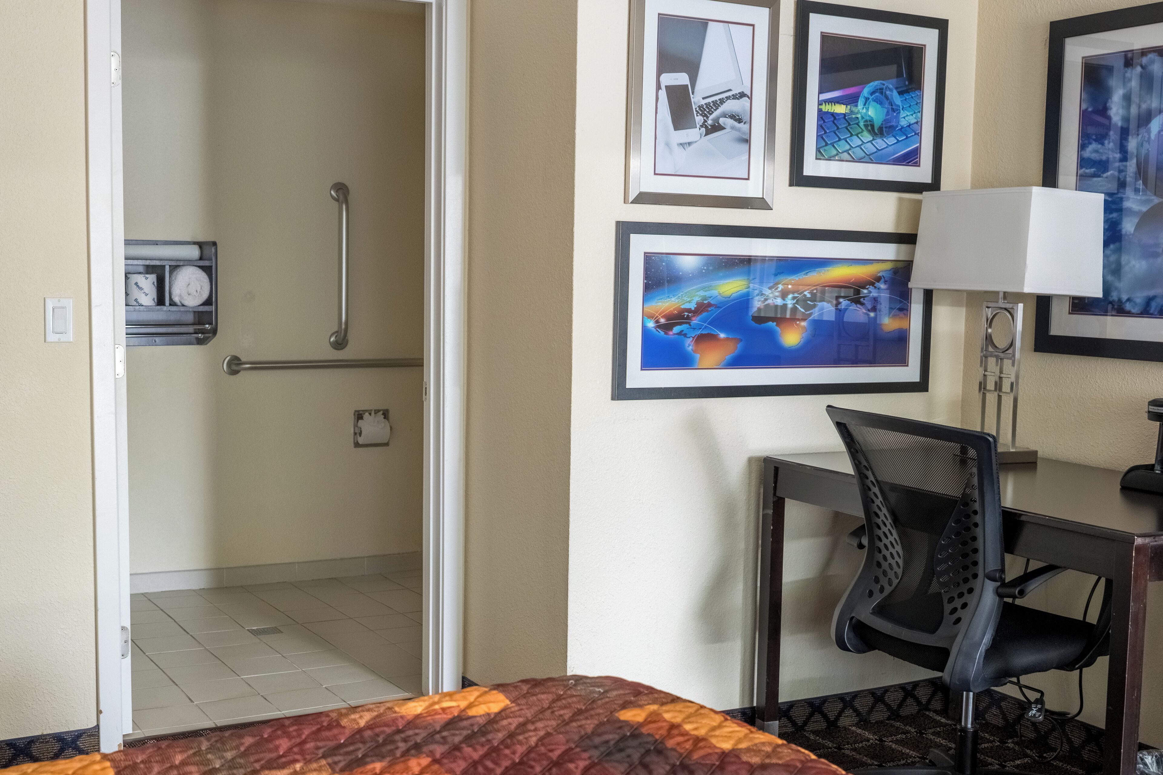 A hotel room with a bed, a desk with a chair, a desk lamp, a wall-mounted TV, and a door leading to a bathroom.