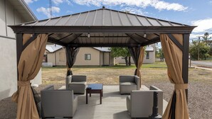 A covered outdoor seating area with four chairs and a table, a grill, and a pergola with curtains. - Oregon Trail Inn and Suites (Lakeview)