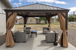 Un área de estar al aire libre techada, con cuatro sillas y una mesa, una parrilla y una pérgola con cortinas.