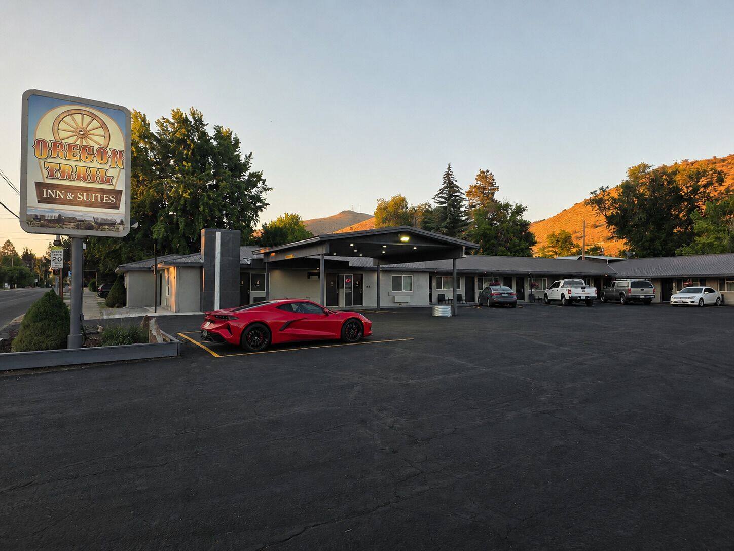 A motel with a sign for Oregon Trail Inn & Suites, a parking lot, and a red sports car parked in front.
