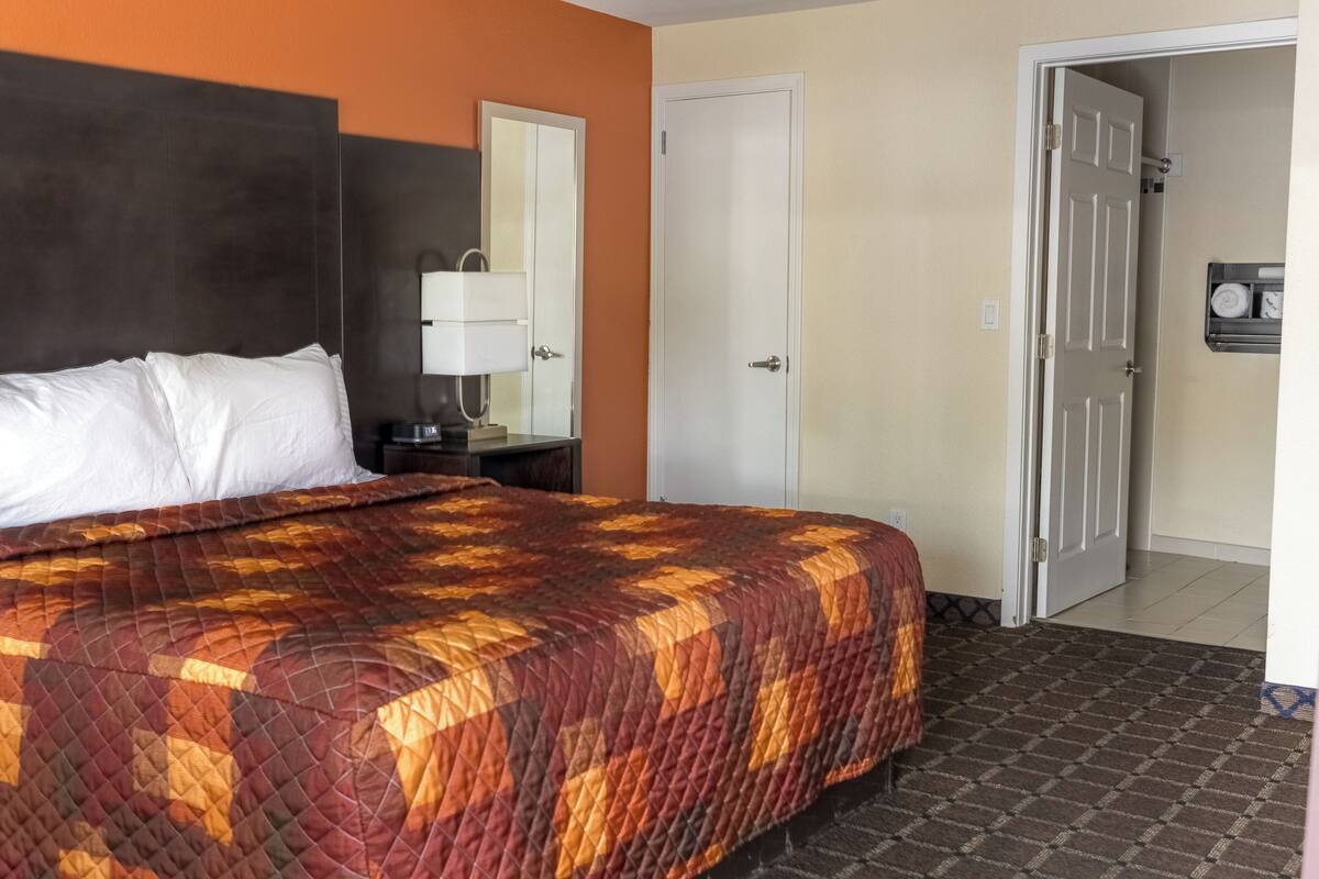A hotel room with a bed, a nightstand, a lamp, a door, and a wall-mounted shelf with towels.