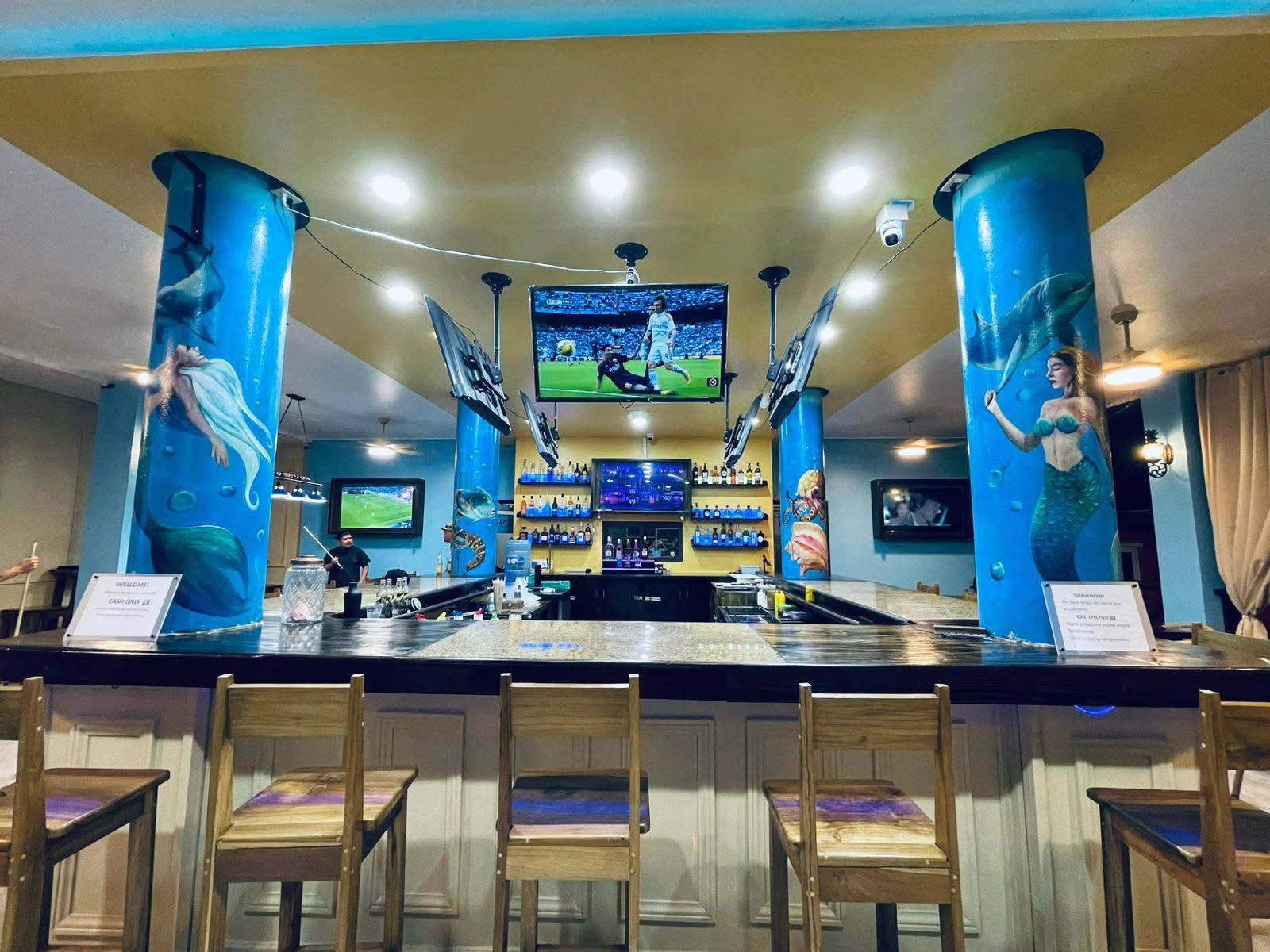 A bar area with multiple screens, a bar counter, and wooden chairs.