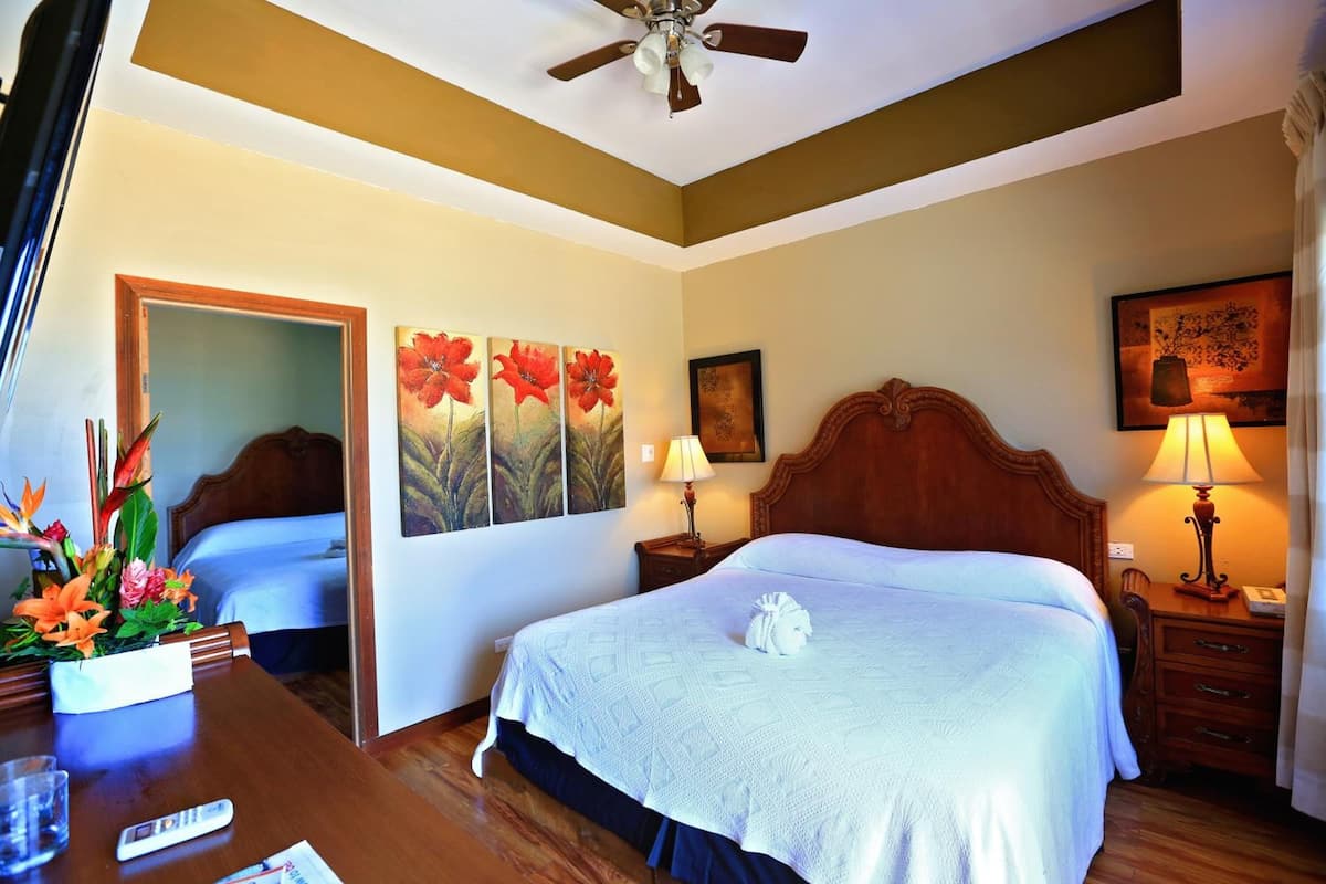 A hotel room with a bed, bedside tables, a desk with a lamp, a ceiling fan, and artwork on the walls.