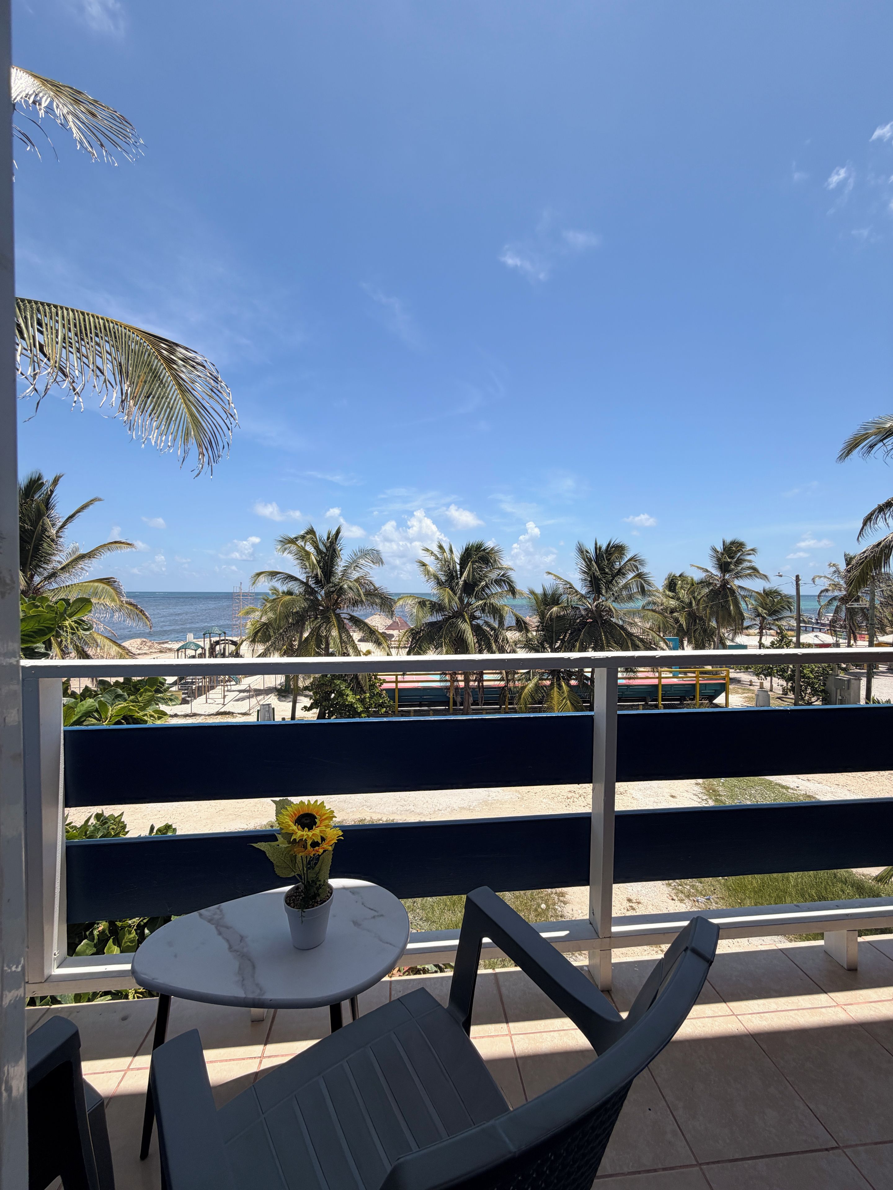 A balcony with a table and chairs, overlooking a beach with palm trees and a clear sky.