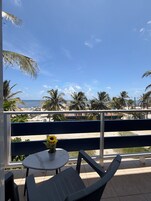 Een balkon met een tafel en stoelen, met uitzicht op een strand met palmbomen en een heldere lucht.