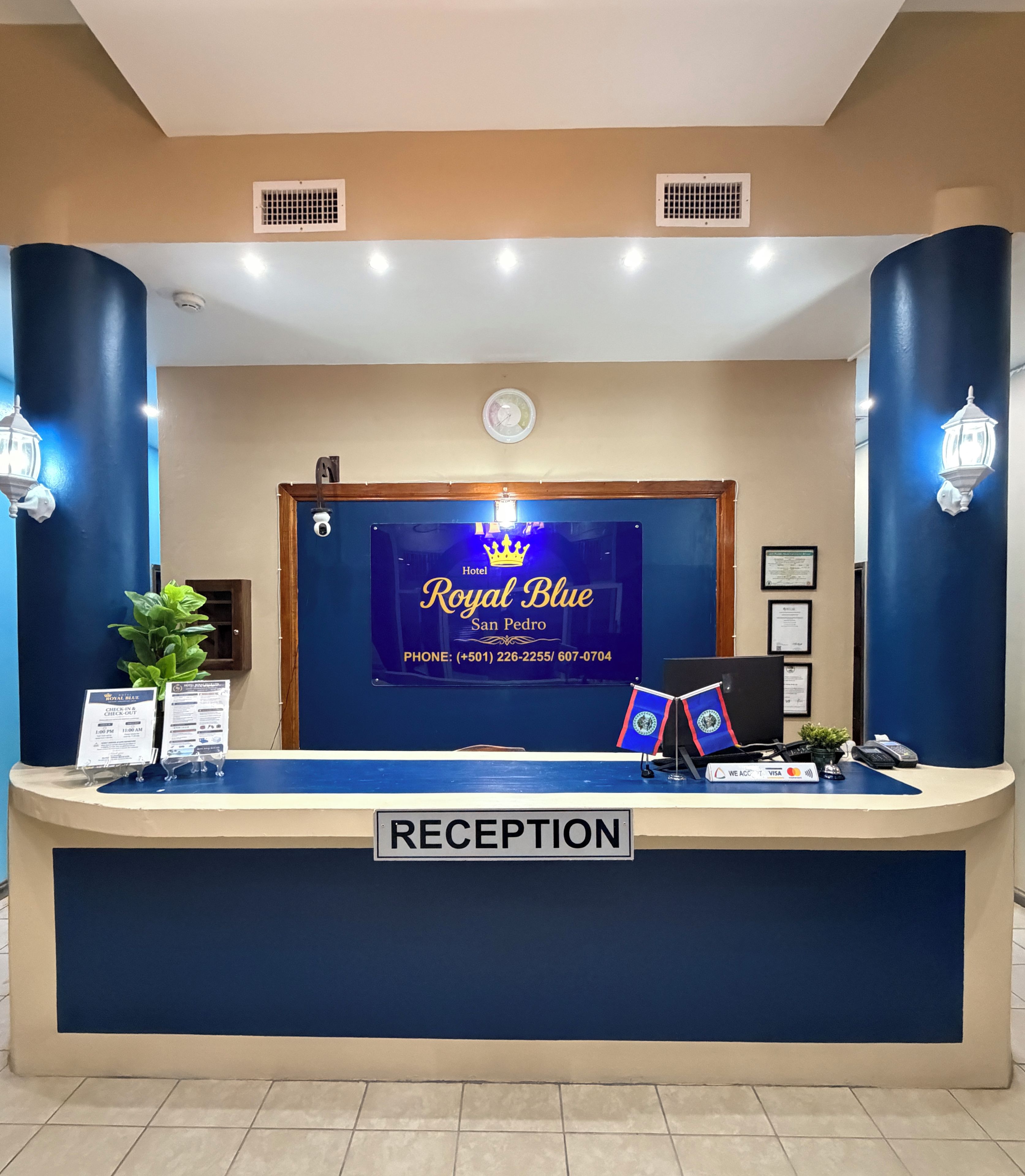 Mostrador de recepción en el Hotel Royal Blue con un cartel que indica horarios de registro de entrada y salida, un teléfono y una planta.
