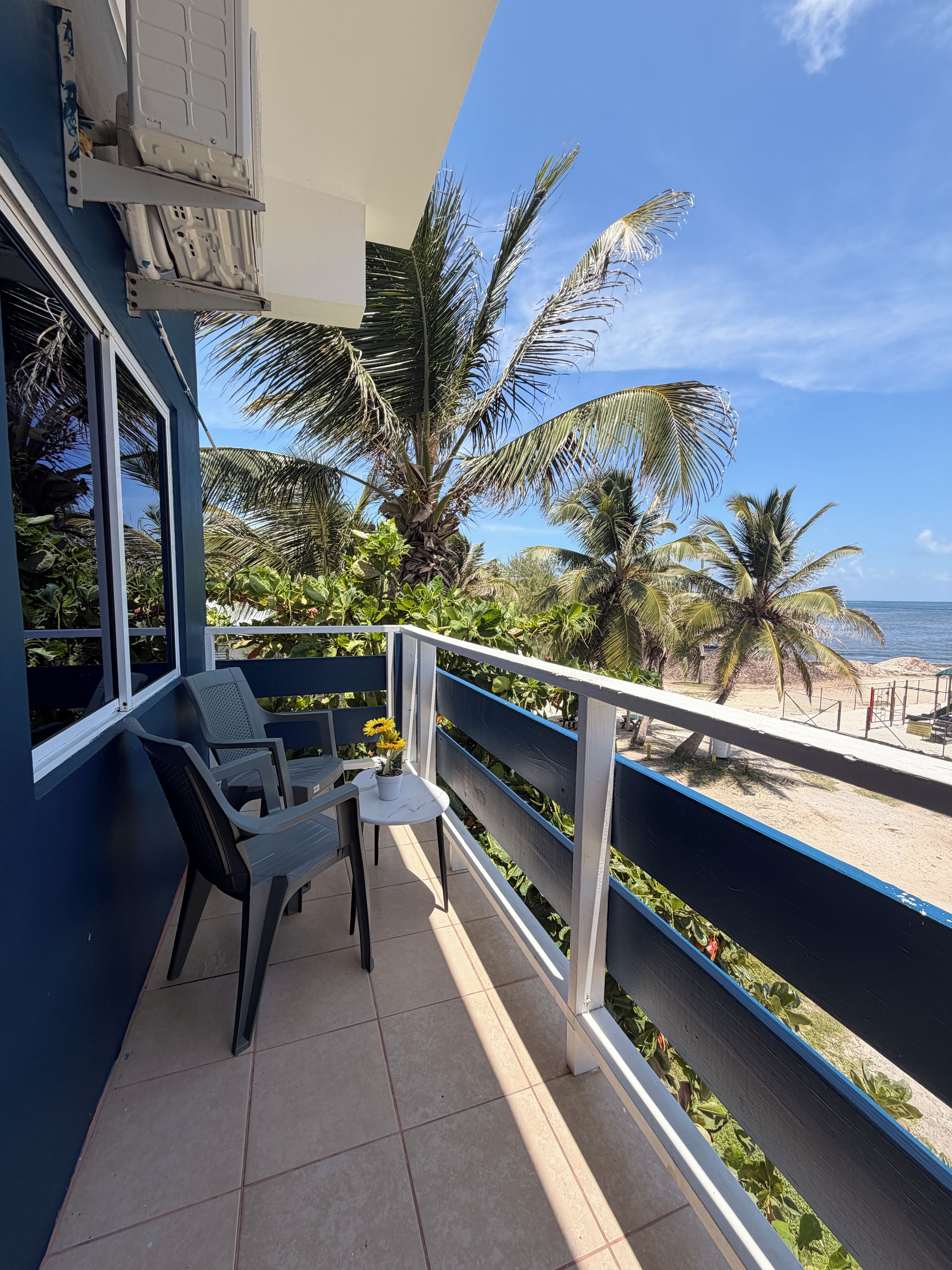A balcony with a view of the beach, equipped with a table and chairs, and surrounded by palm trees.