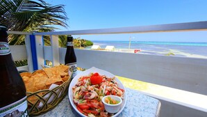 A table with a shrimp salad, chips, and a bottle of beer, overlooking a beach.