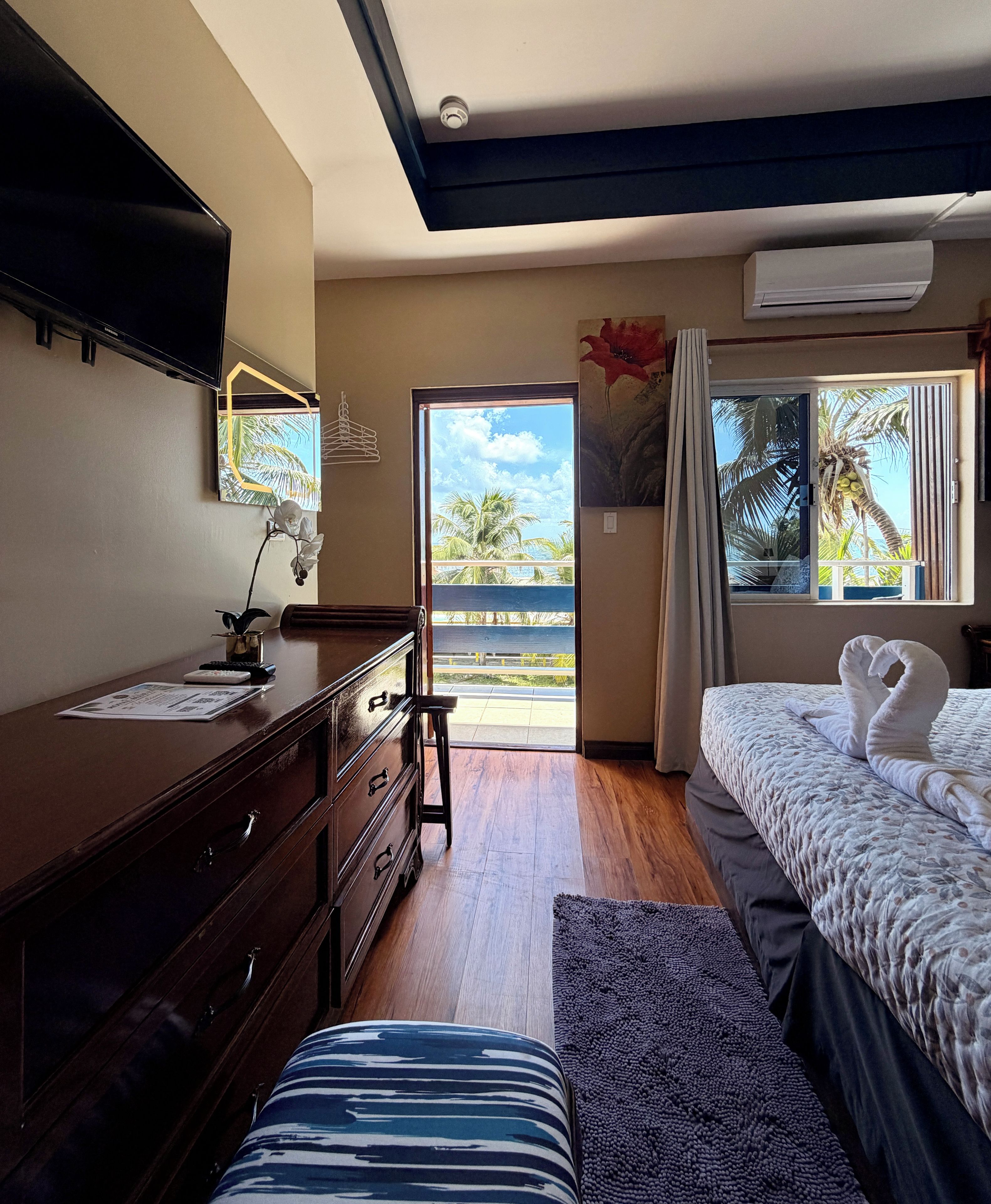 Una habitación de hotel con una cama, un escritorio con computadora, un aparador, una ventana con vista a palmeras y un equipo de aire acondicionado.