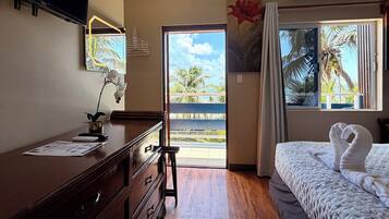Una habitación de hotel con una cama, un escritorio con computadora, un aparador, una ventana con vista a palmeras y un equipo de aire acondicionado.