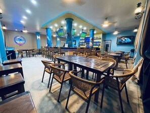 A restaurant interior with multiple dining tables and chairs, a bar area, and a television screen mounted on the wall. - Hotel Royal Blue & Golf Cart Rental (San Pedro)