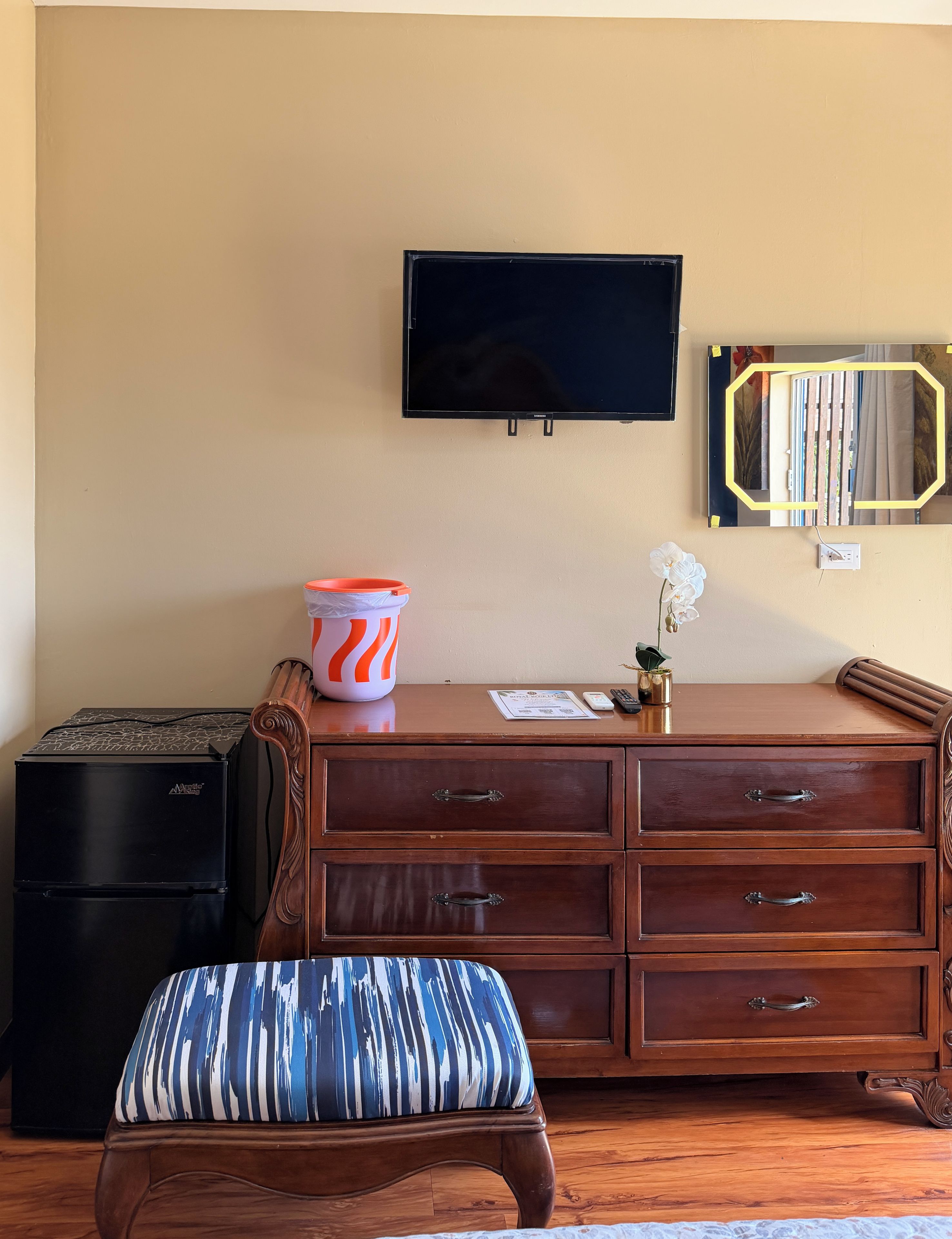 Una habitación con un aparador de madera, un televisor montado en la pared, una mesita con un florero y un banco.