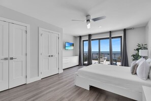 A modern bedroom with a large bed, a ceiling fan, a TV, and a balcony with a view of the sea.