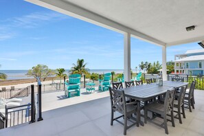 Un espacio al aire libre techado con vista a la playa, que incluye una mesa, sillas y una barandilla.