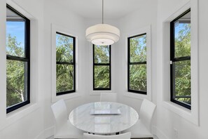 Une pièce avec une table ronde et quatre chaises, de grandes fenêtres à encadrements noirs et un luminaire au plafond.