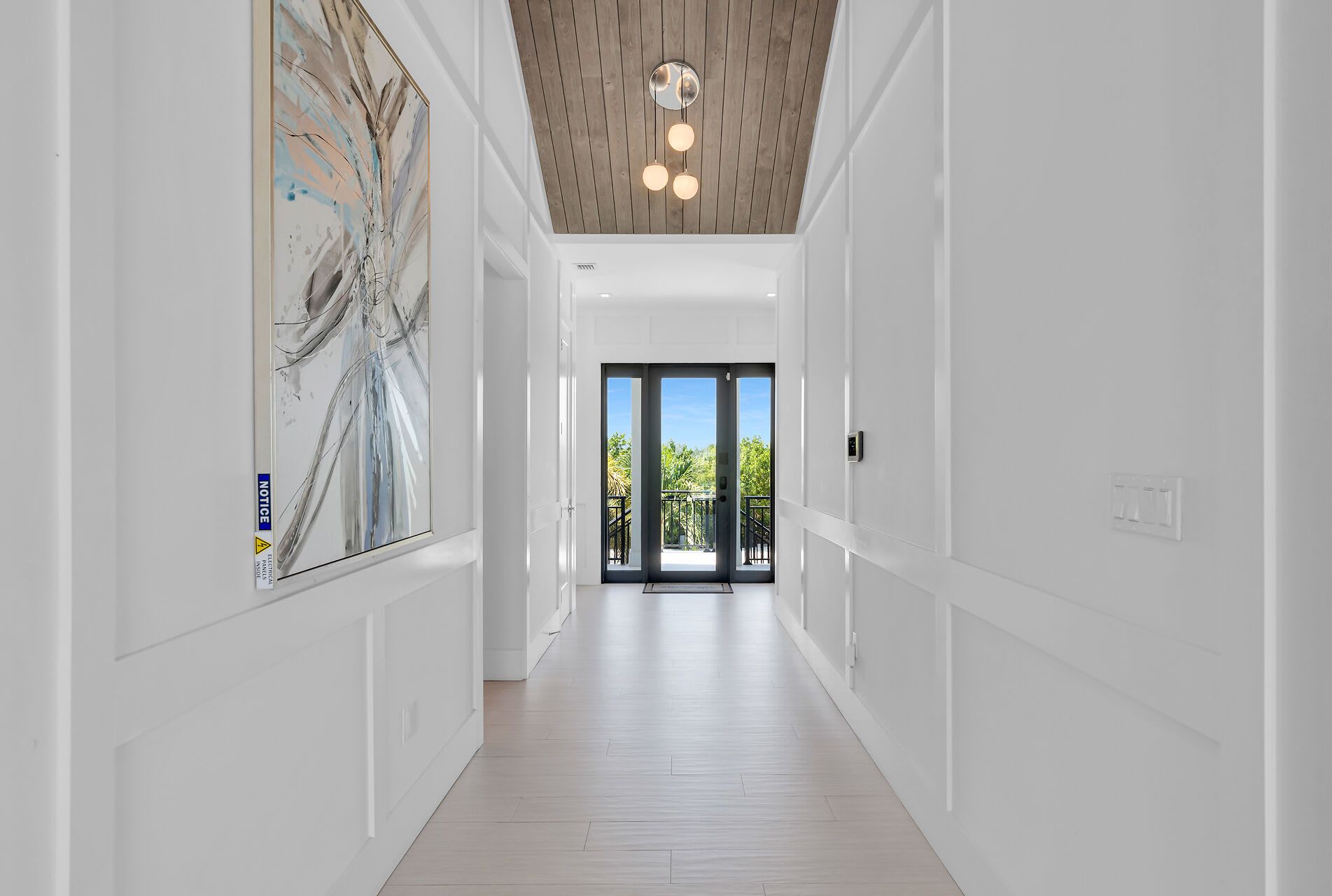 Un couloir avec un plafond en bois, une grande peinture abstraite sur le mur de gauche et, au fond, une vue sur un balcon agrémenté de verdure.