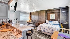 Una cocina compacta y moderna con encimera de mármol y taburetes, una zona de estar acogedora con sofá y una mesita, y una cama grande con colcha estampada.